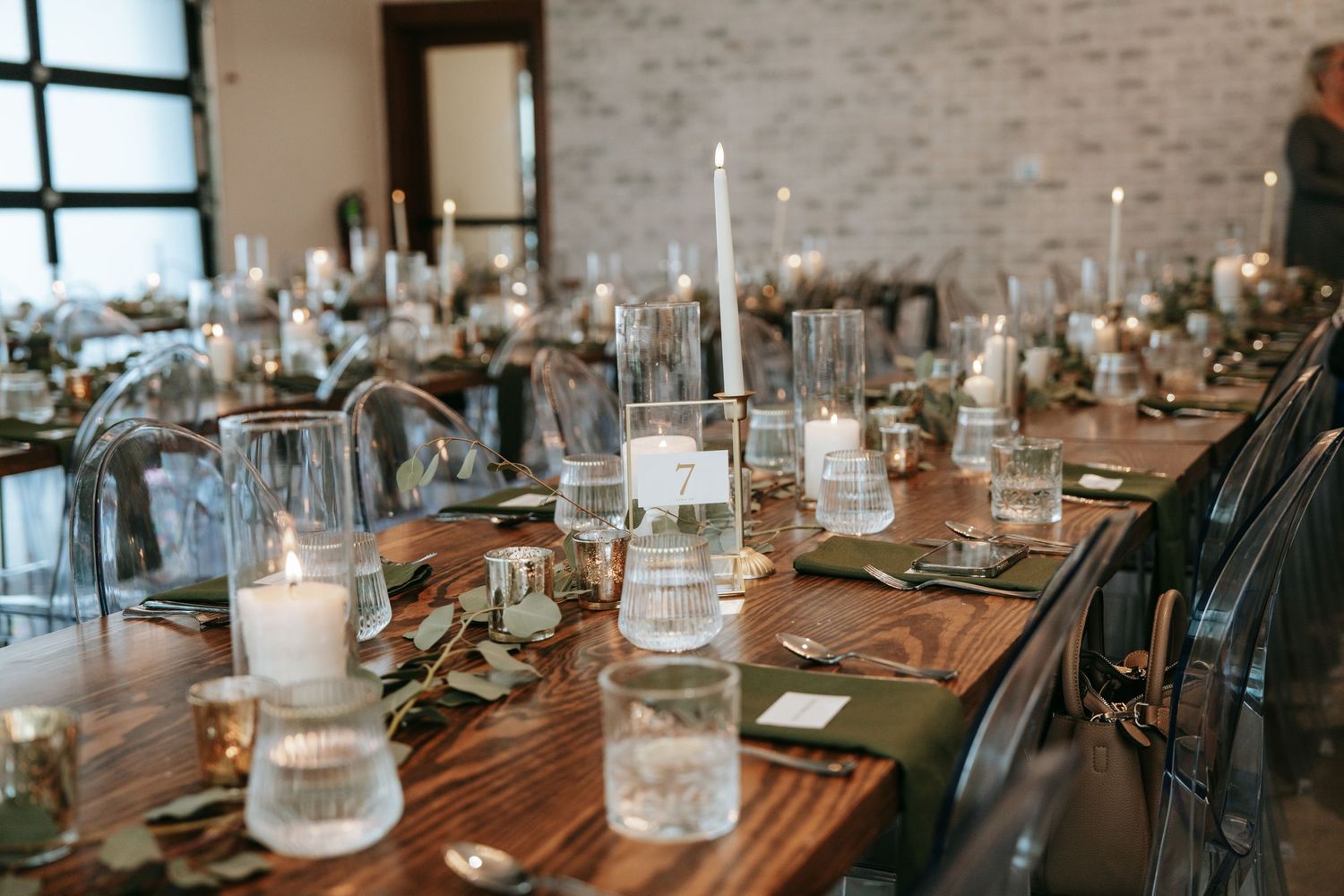 Rustic wedding reception tables with candles, greenery, and glass centerpieces on dark wood tables.
