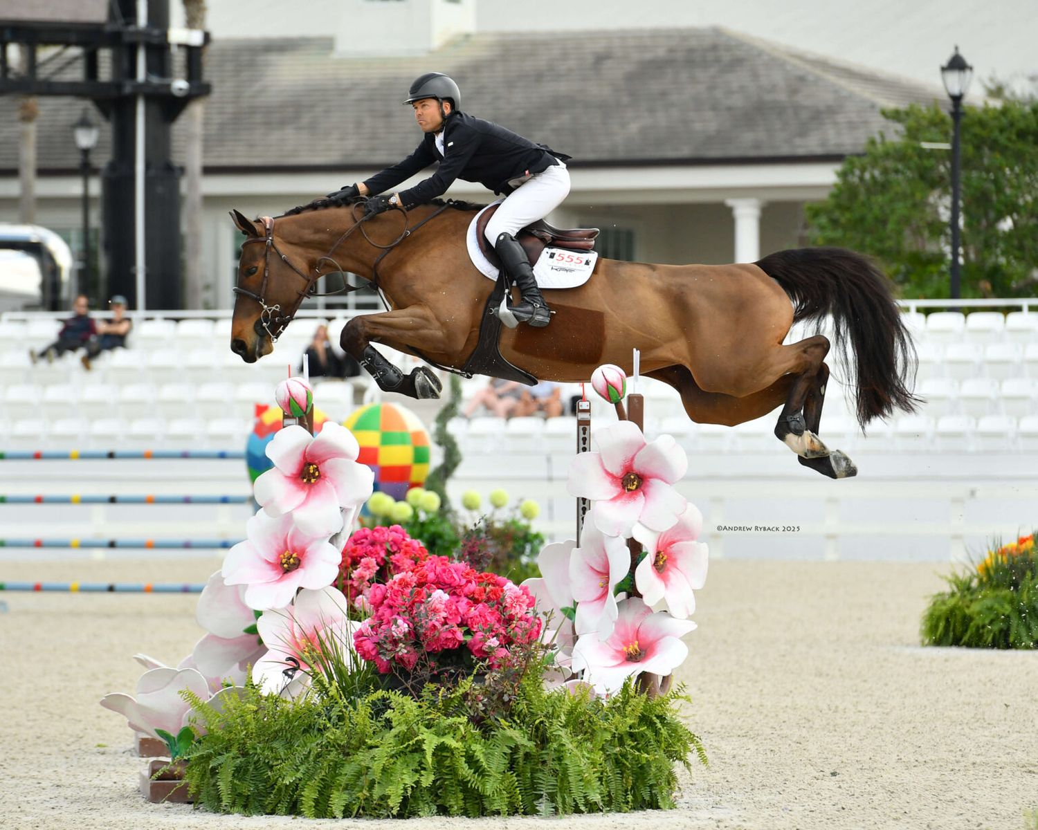 Show jumping horse and rider clearing a floral jump obstacle at an equestrian competition.