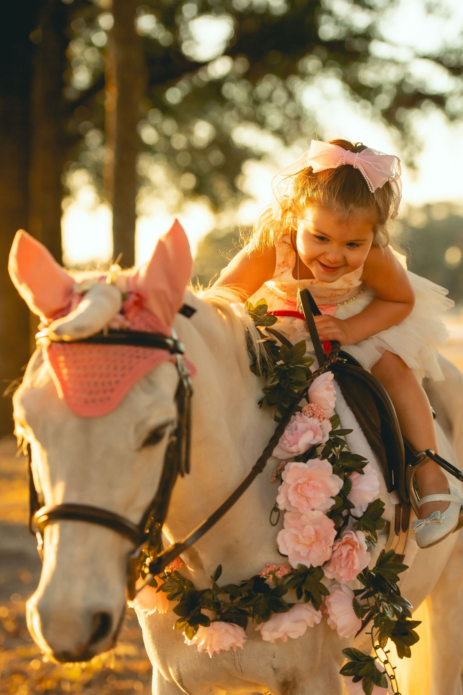 White horse decorated with pink roses carries a small rider in a white dress at sunset.