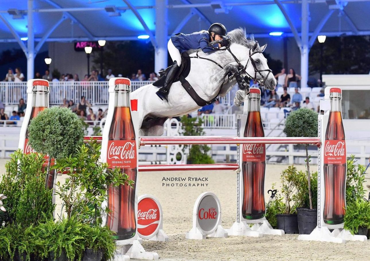 Show jumping competition with Coca-Cola themed obstacles in illuminated arena setting.