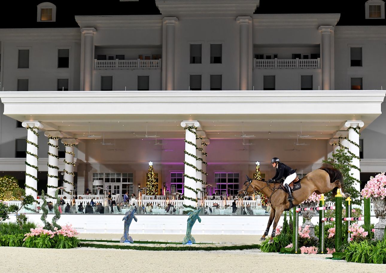Ice skaters glide across an illuminated outdoor rink beneath striped columns and palm trees at night.