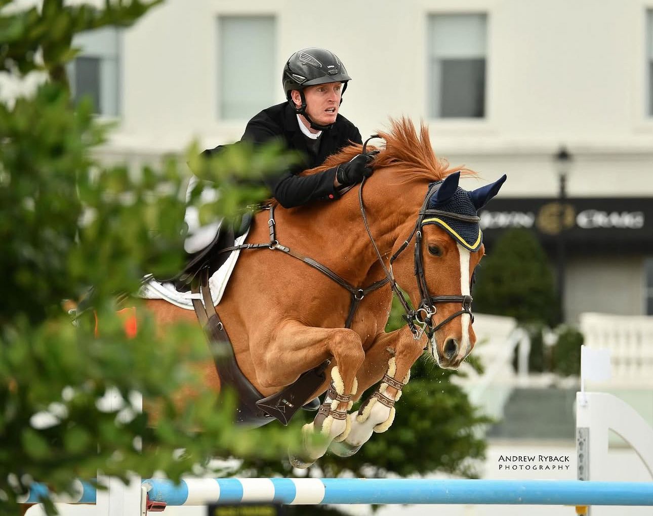 Chestnut horse and rider soaring over blue jump rail during equestrian competition.