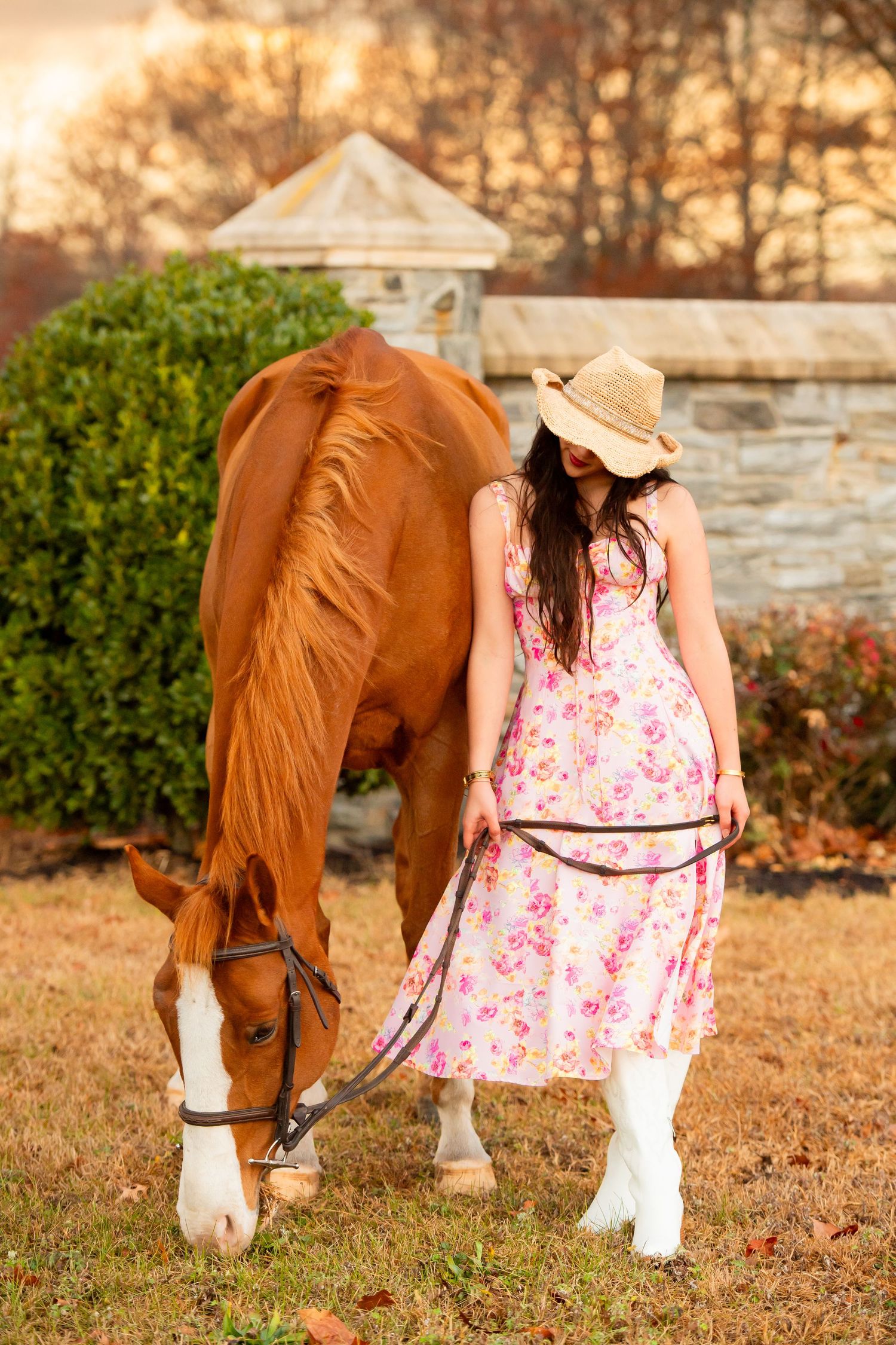 Chestnut horse grazing while companion in floral dress and boots stands nearby.