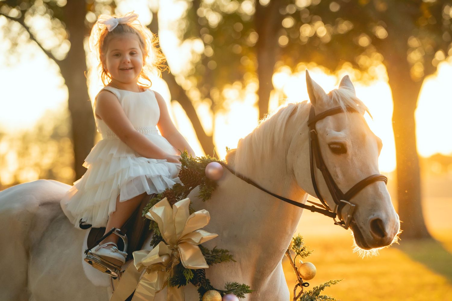 Child in white dress rides decorated white pony at sunset through trees.