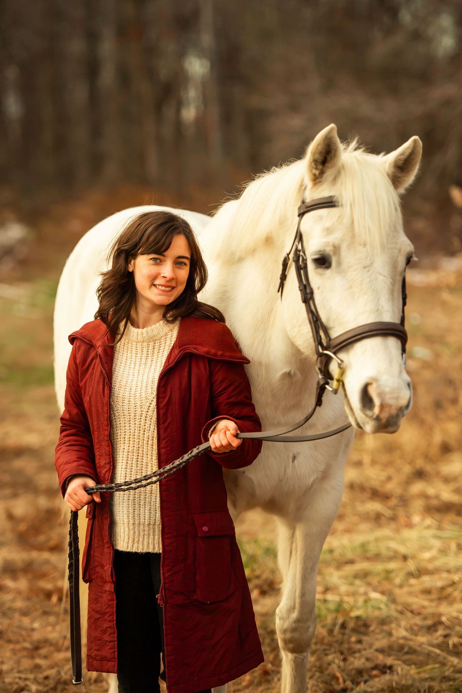 A majestic white horse with bridle stands next to a person wearing a red coat and cream sweater in a fall setting.