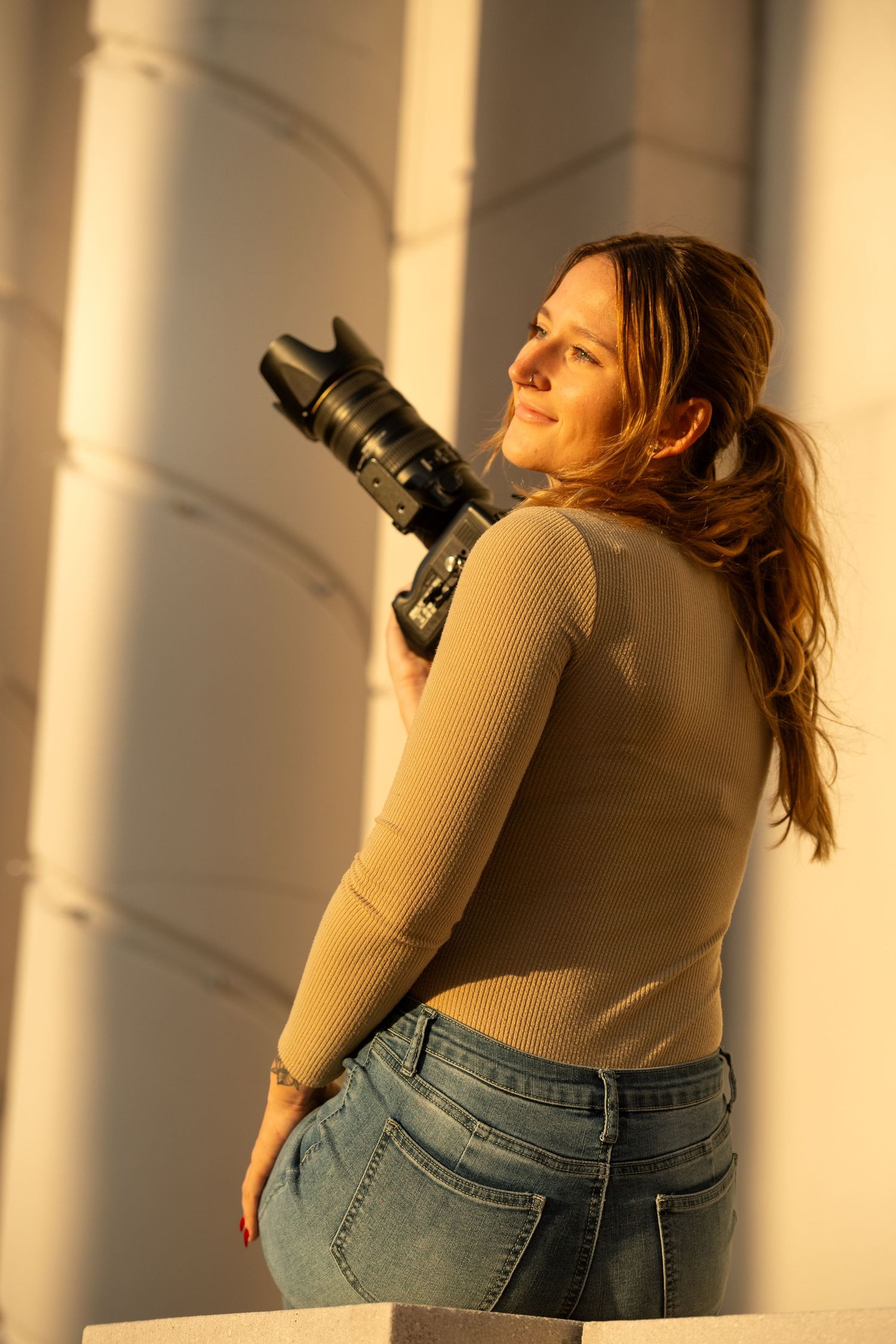 Someone holding professional camera equipment poses against white columns in golden sunset light.