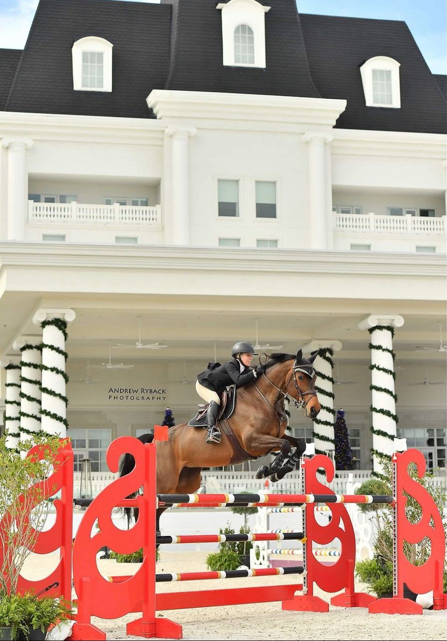 Horse and rider jumping over red obstacles in front of a grand white colonial-style building.
