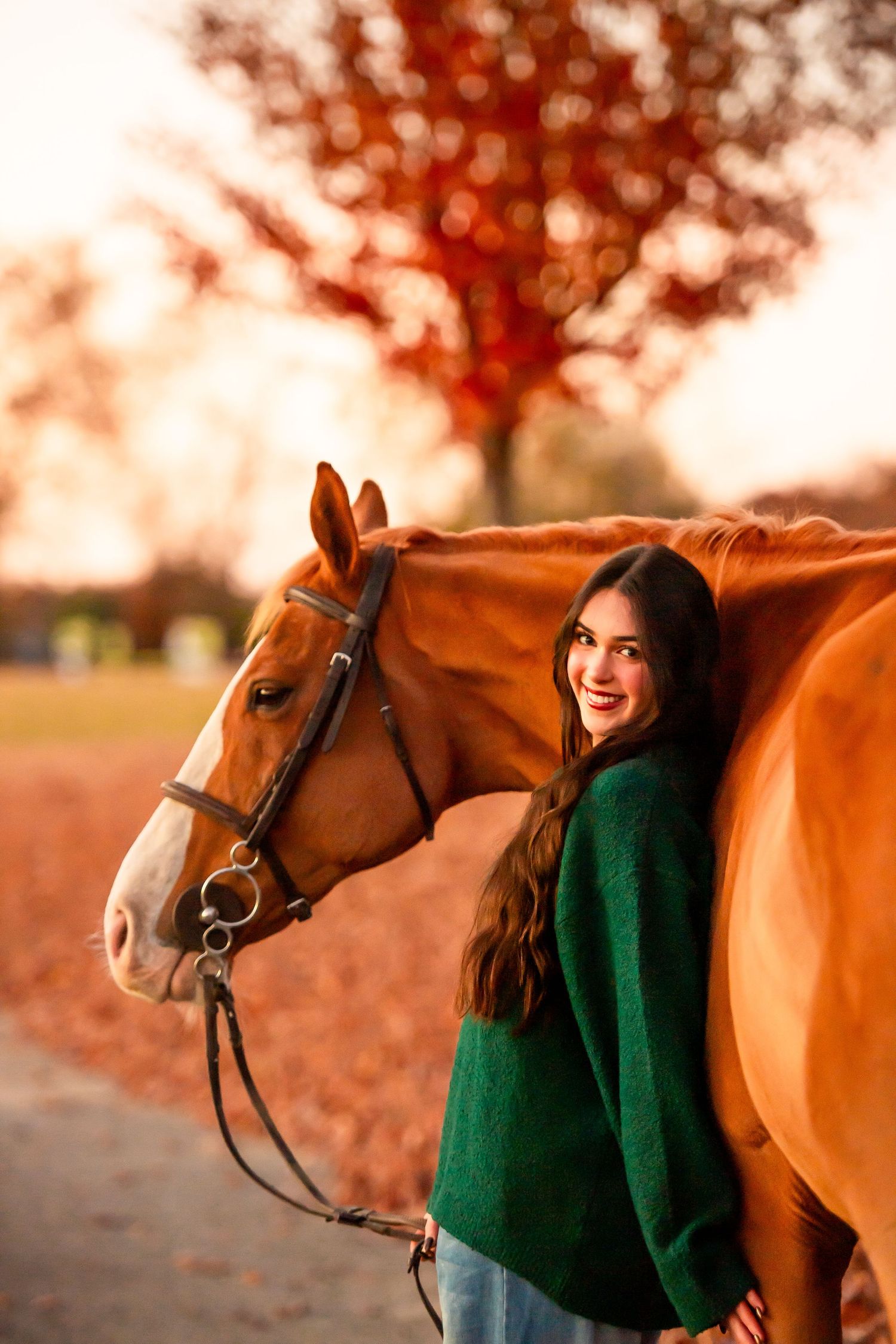 Chestnut horse and person in green sweater pose together during golden hour with red autumn trees.