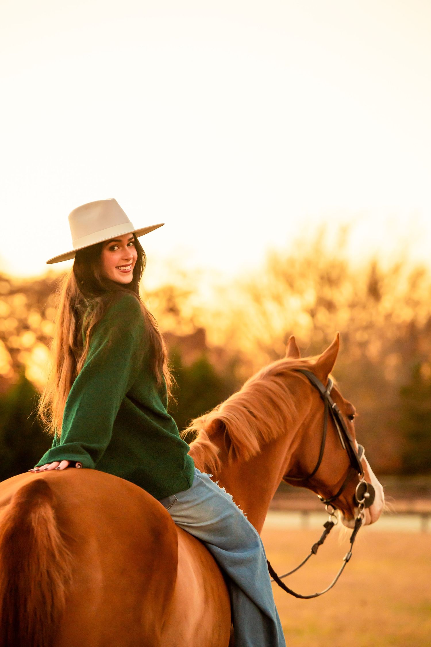 Equestrian riding a chestnut horse at sunset wearing green sweater and tan hat.