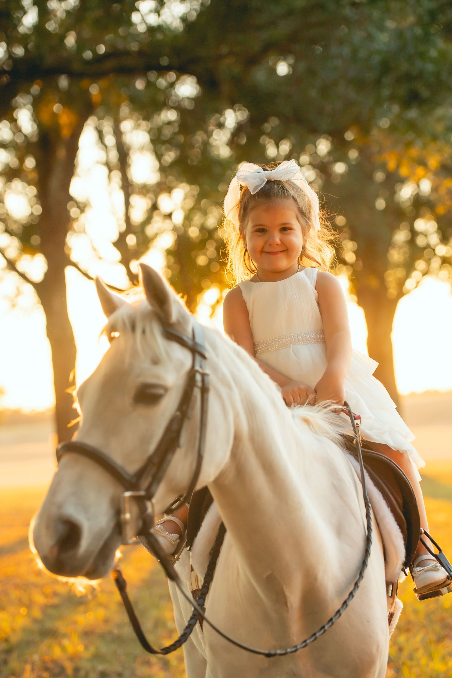 Child in white dress rides white horse during golden hour.