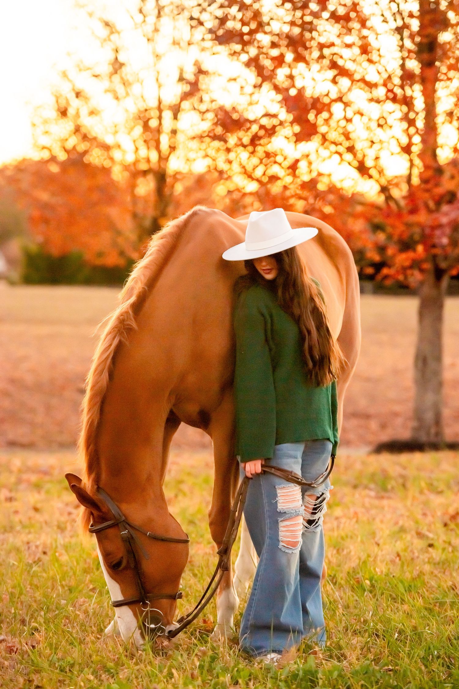 A chestnut horse grazes while a person in western wear stands nearby during autumn sunset.