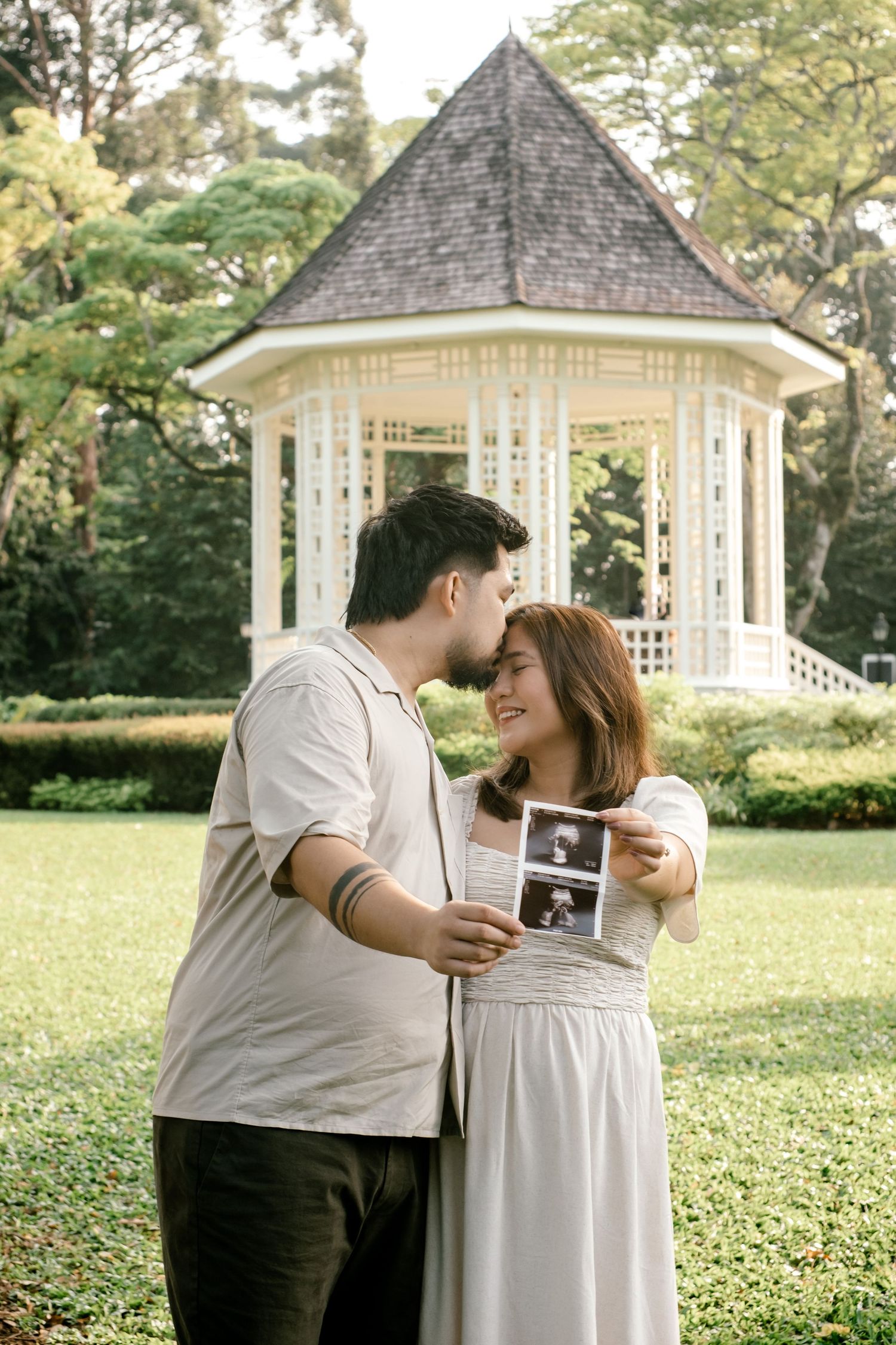 Couple shares a tender kiss while holding ultrasound photos in front of a bandstand, Botanic Garden.