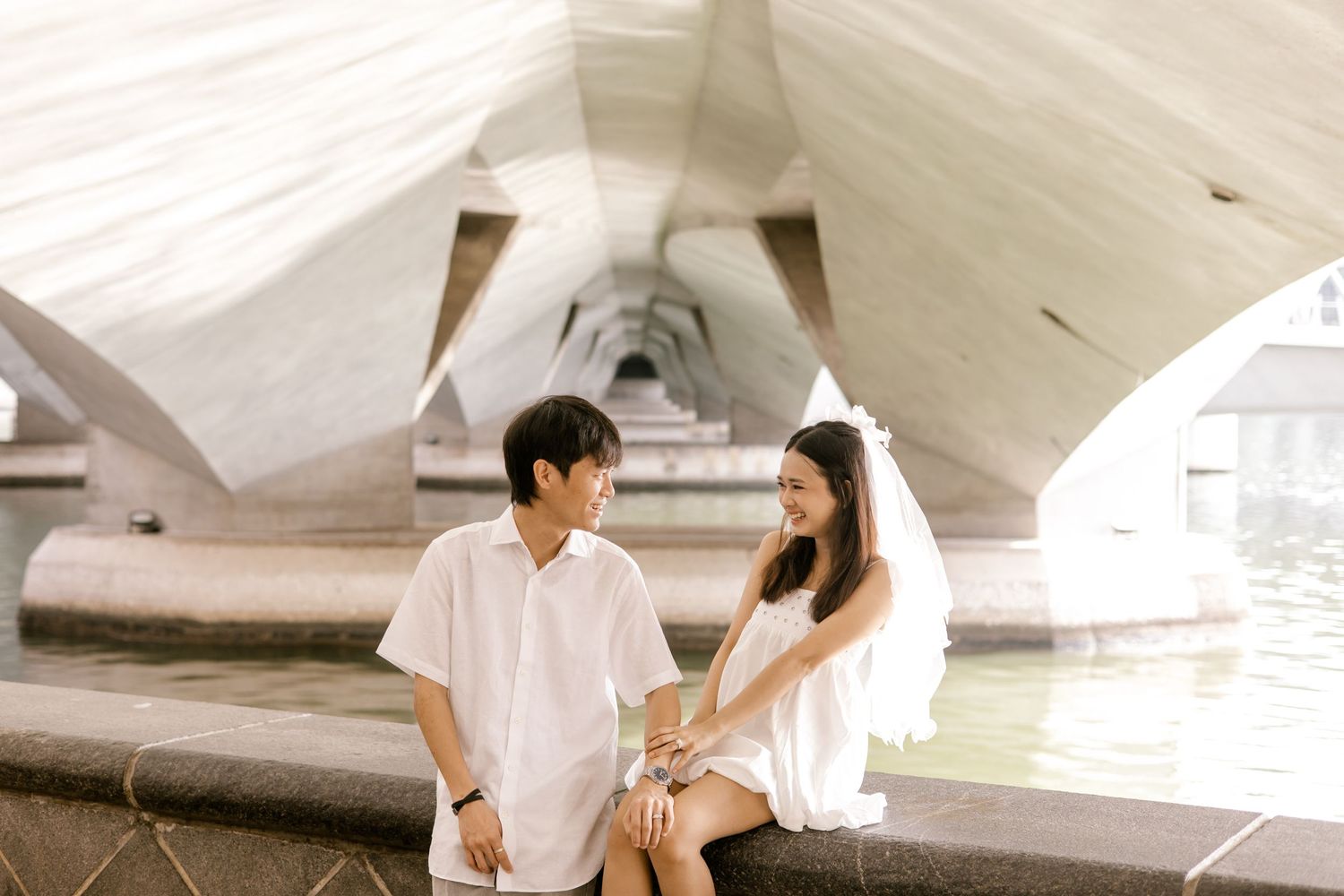 A young Asian couple sits by a waterway under a modern white architectural structure, sharing a tender moment.
