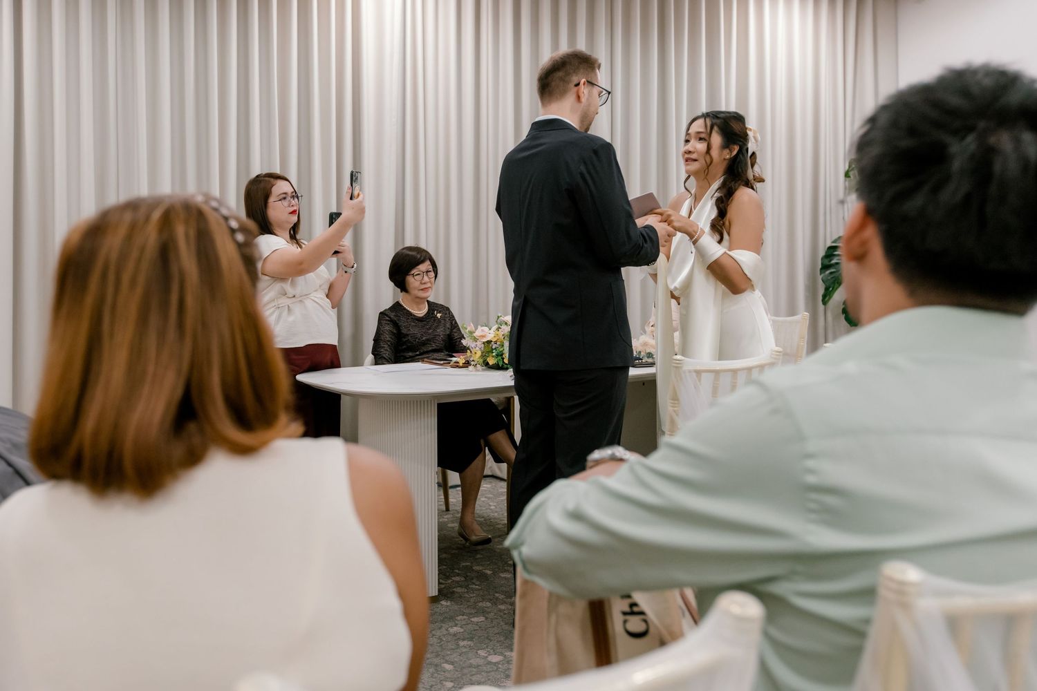 Couple exchanging vows at intimate wedding ceremony with guests seated and woman capturing moment on phone.