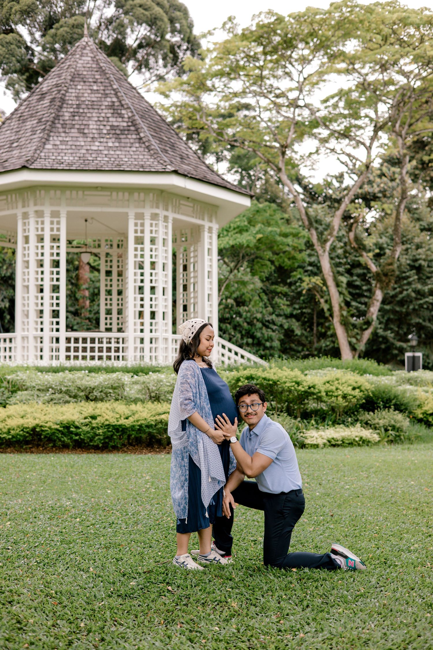 Pregnant couple posing in a lush park with a white gazebo, man kneeling to touch baby bump.