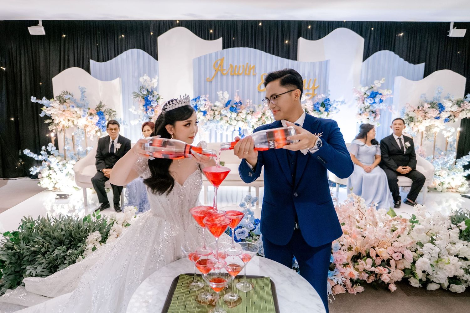 Bride and groom pouring champagne tower at elegant wedding reception with floral decorations and blue backdrop.
