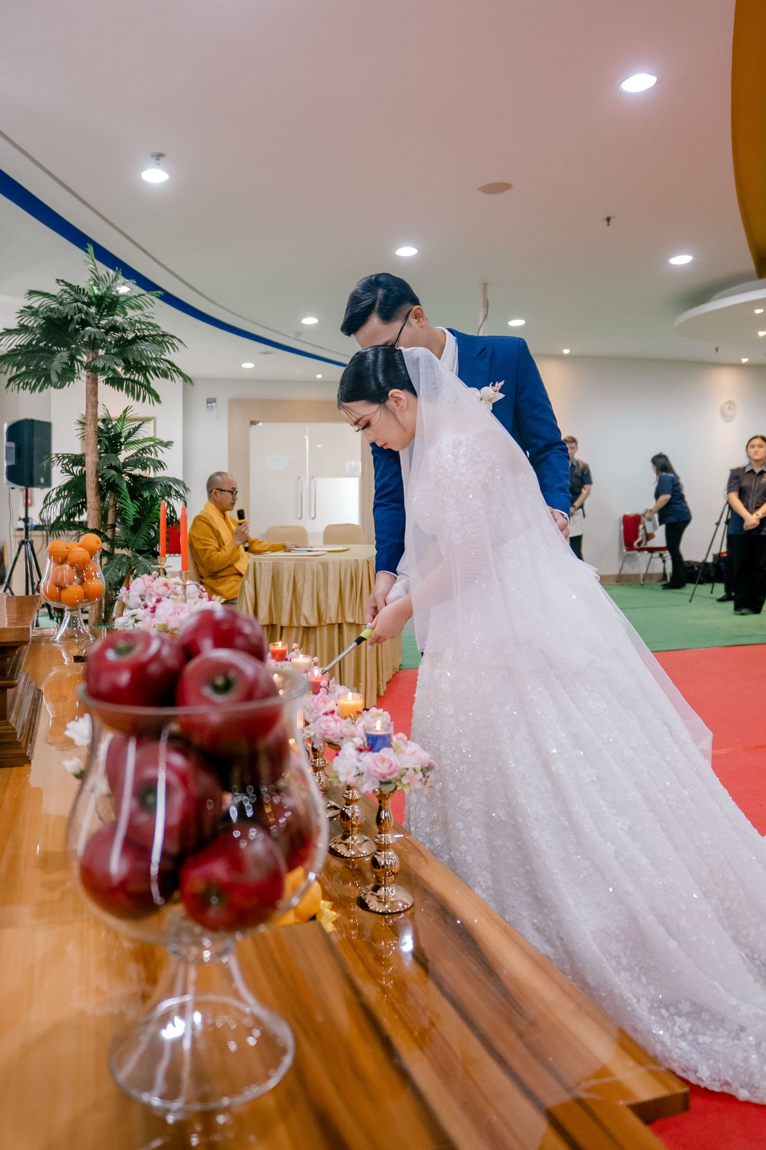 Bride and groom lighting candles at a Buddhist wedding ceremony with red apples and floral offerings on altar.