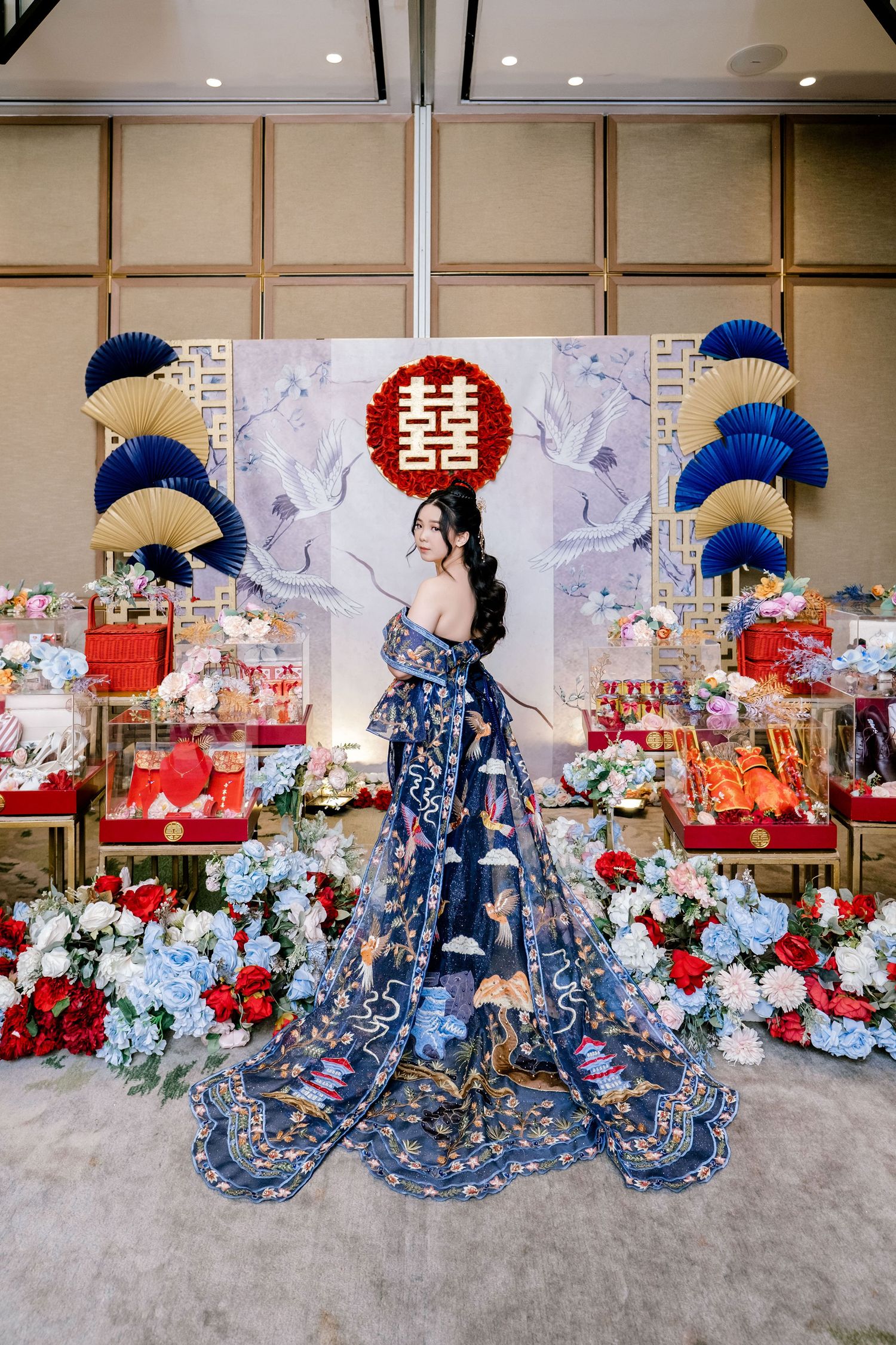 Woman in ornate blue floral gown poses at elegant Chinese wedding reception with double happiness symbol decor.