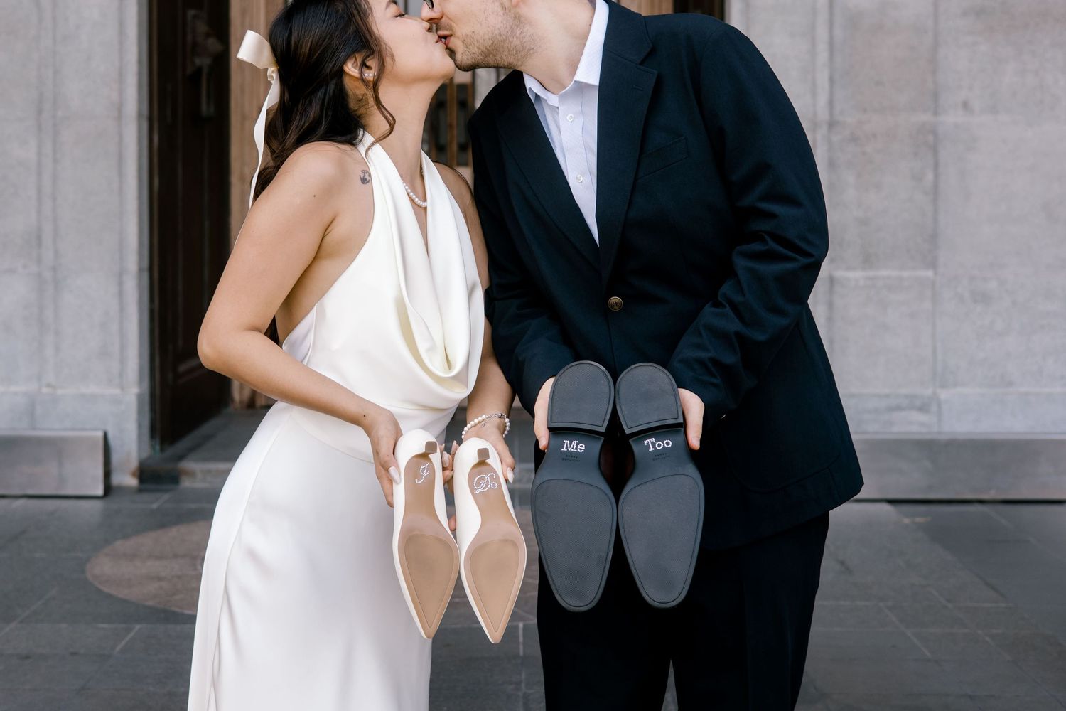 Bride and groom kissing while holding Dior heels and bouquet in elegant wedding attire outdoors.
