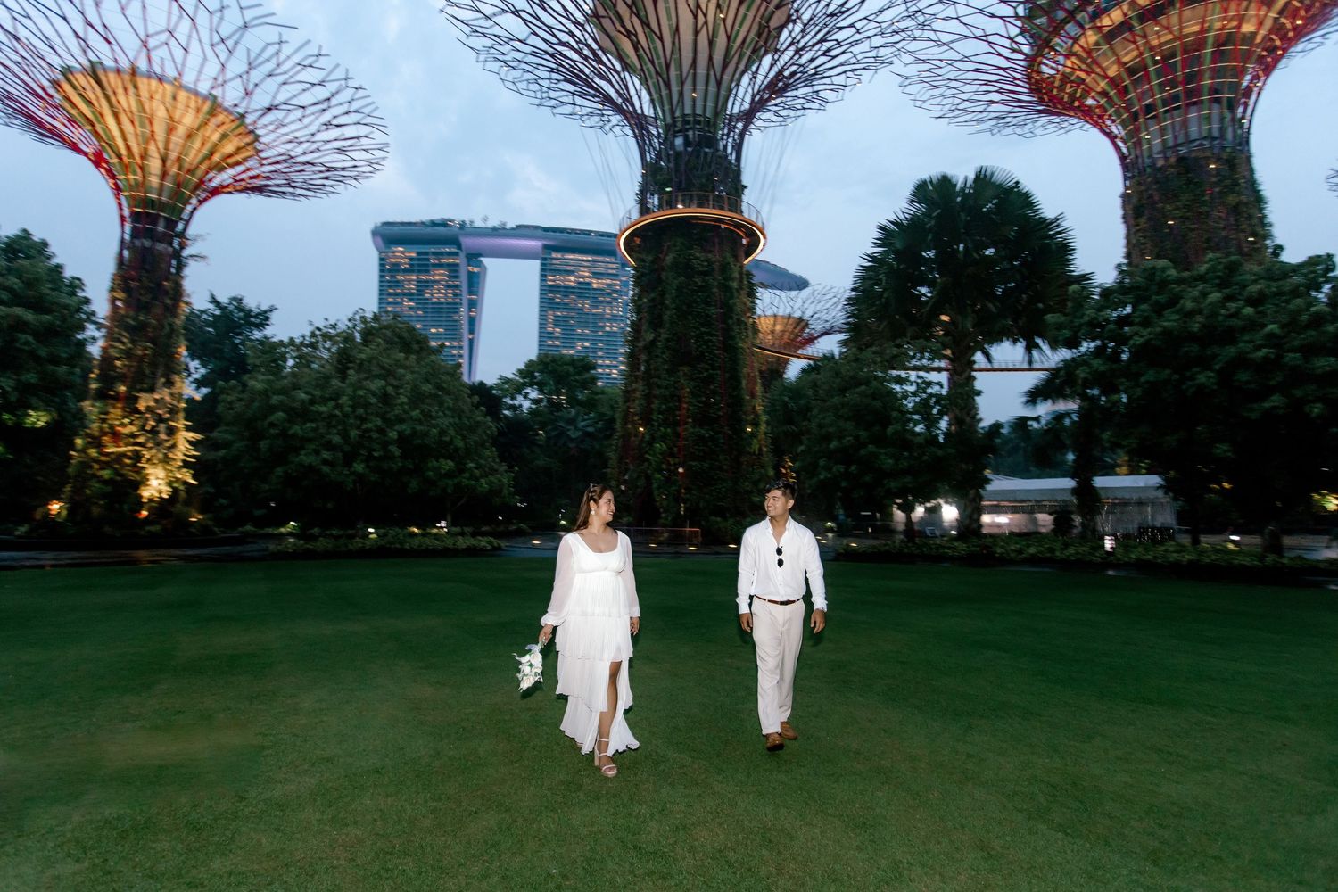 Couple in white attire strolling through Gardens by the Bay, Singapore, with iconic Supertree Grove structures at dusk.
