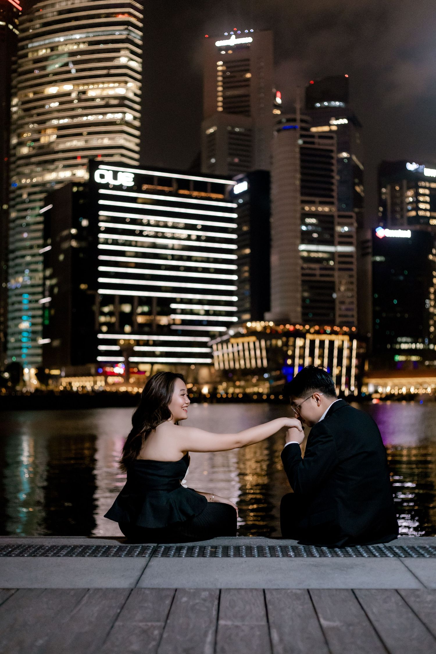 Couple sitting on waterfront at night with illuminated Singapore CBD skyline reflecting on water behind them.