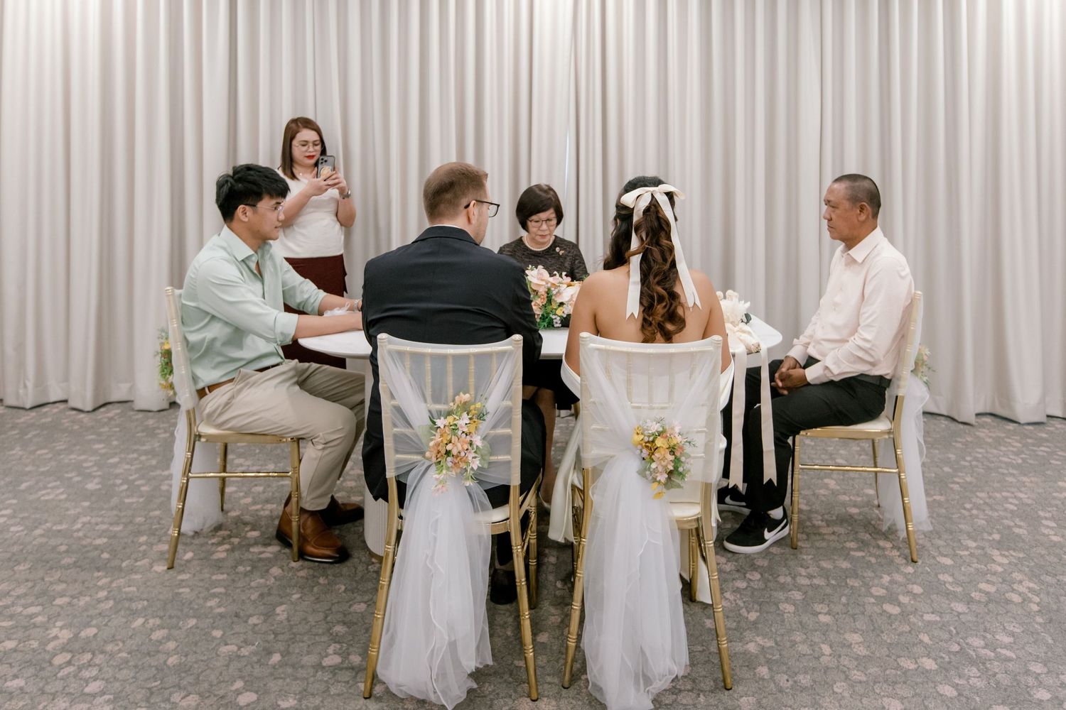 Intimate wedding ceremony with bride and groom signing documents at a decorated table surrounded by family members.