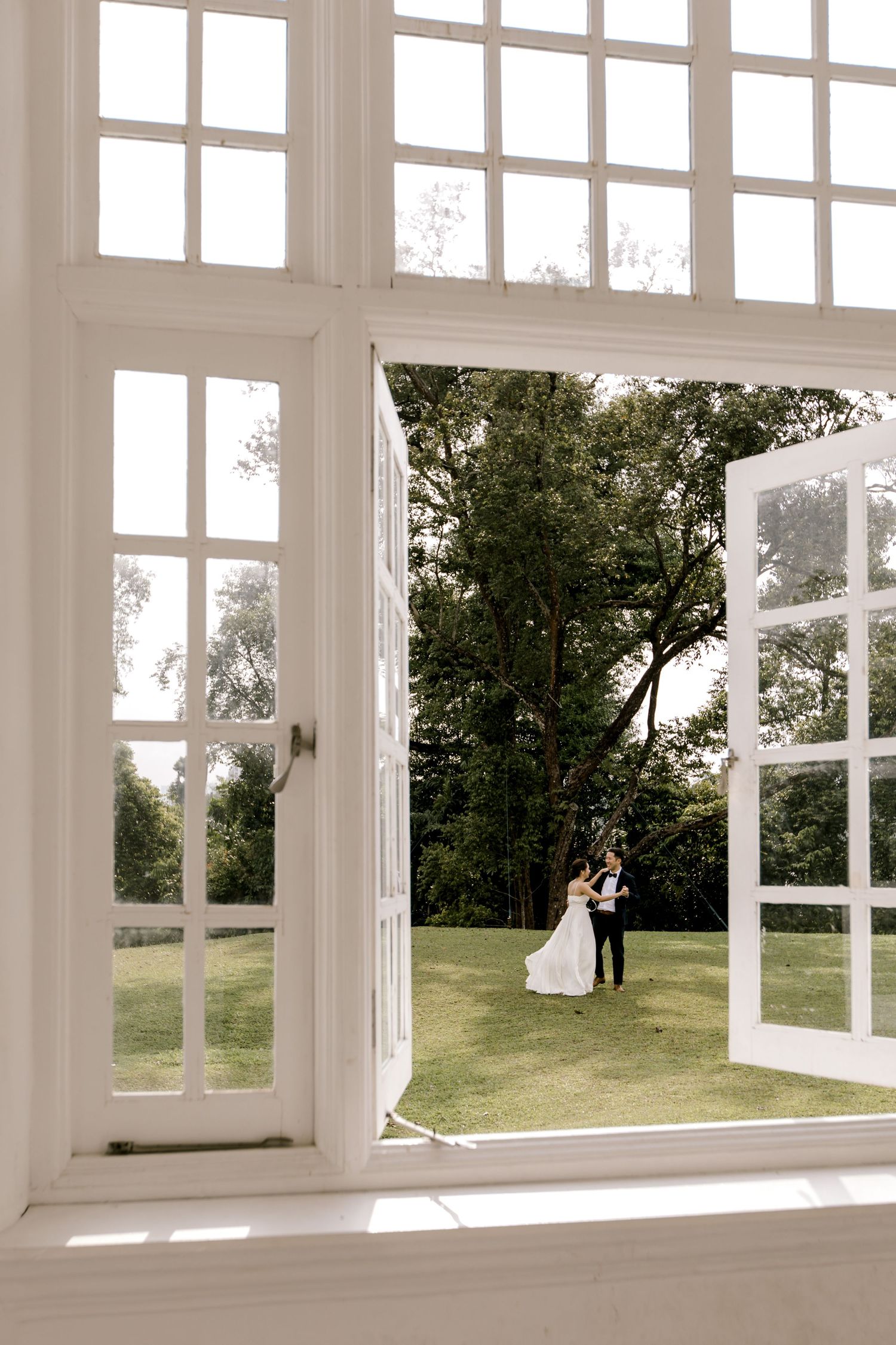 Bride and groom embrace outdoors viewed through elegant white French doors of a bright venue.