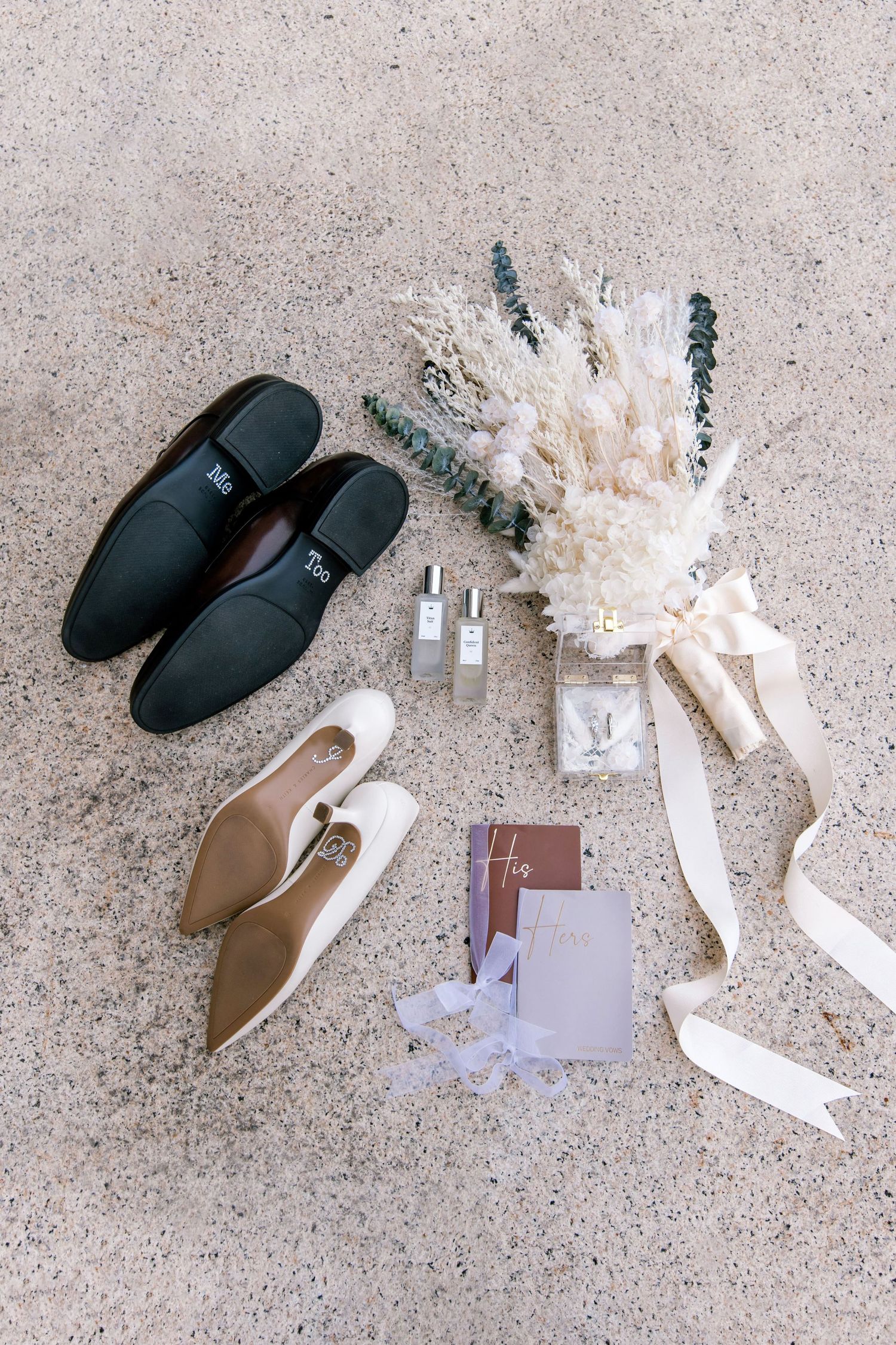 Wedding flat lay featuring his and hers shoes, dried floral bouquet, perfume bottles, rings, and stationery on sandy surface.
