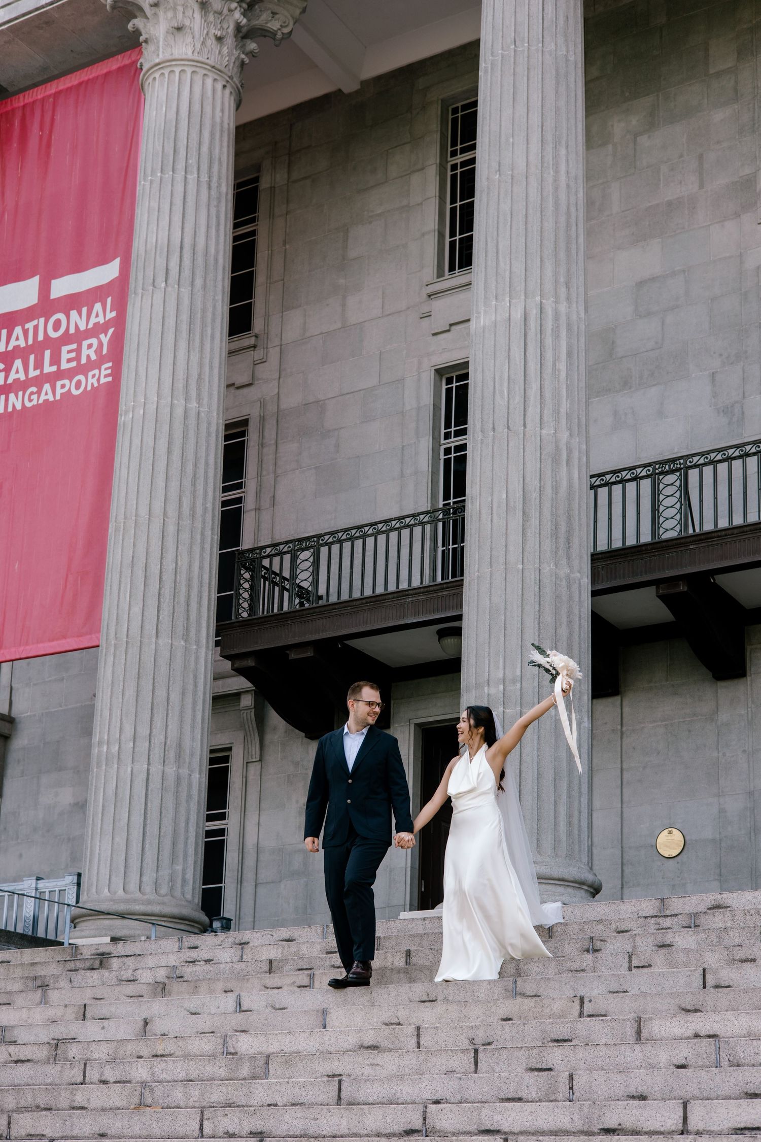 Couple in wedding attire holding hands on steps of National Gallery Singapore, bride raising bouquet joyfully.