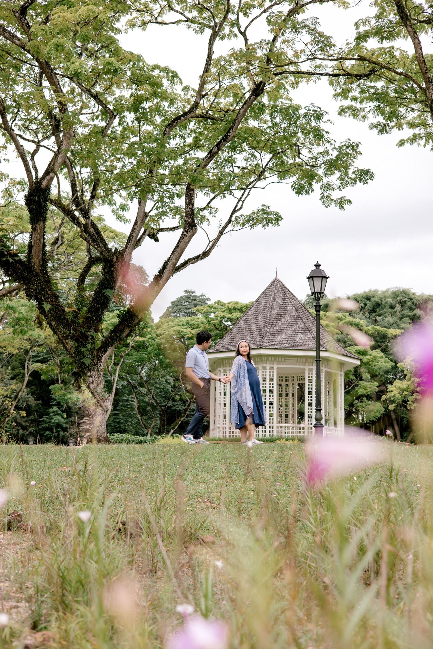 Couple walks hand in hand near a bandstand surrounded by lush greenery and pink flowers.