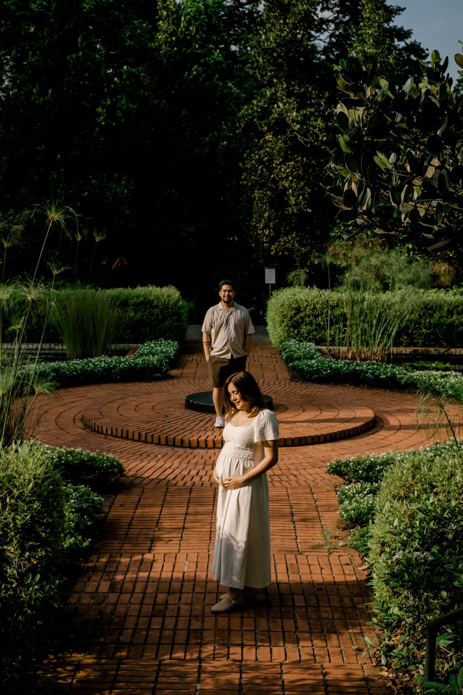 Pregnant woman in white dress cradles her bump in a sundial garden, Singapore with golden hour lighting.