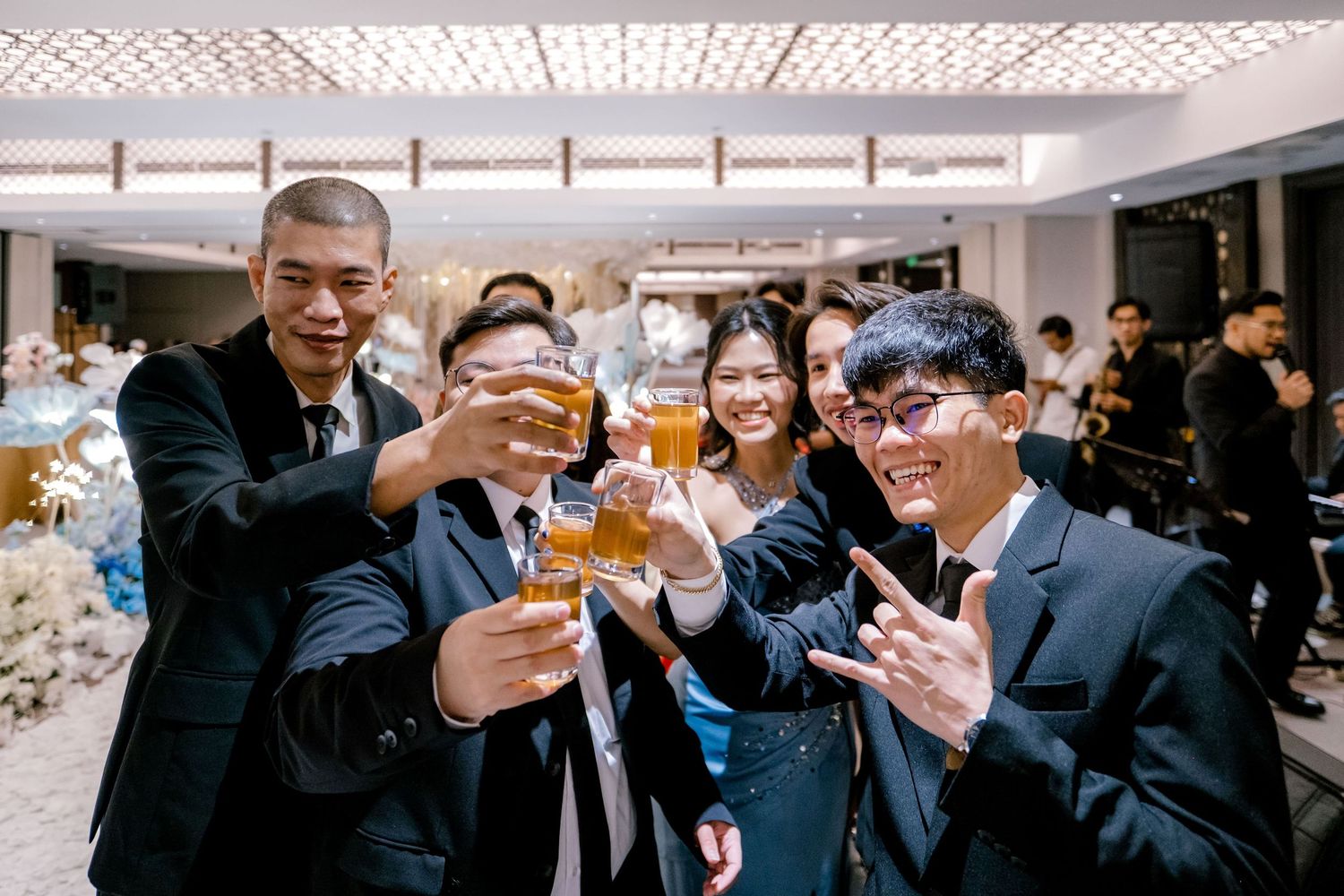 Group of young adults in suits toasting with drinks at a formal wedding reception celebration.