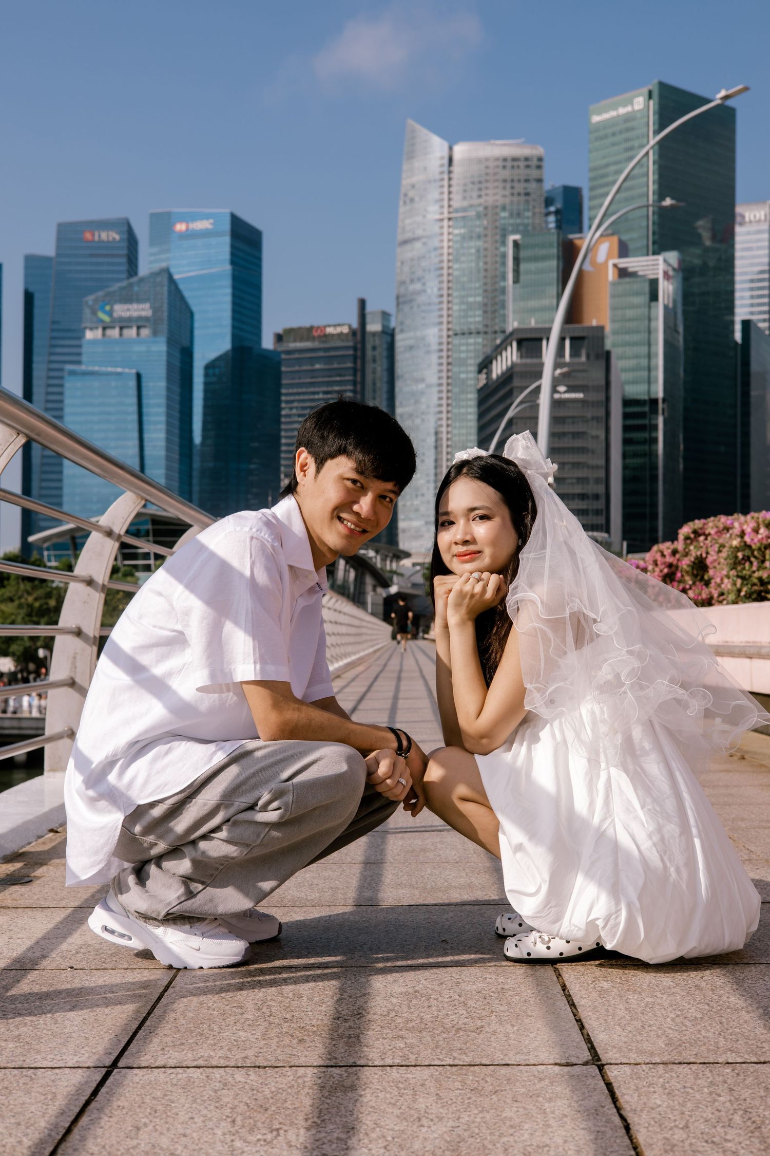 Asian couple in wedding attire posing outdoors with Singapore city skyline in the background on a sunny day.