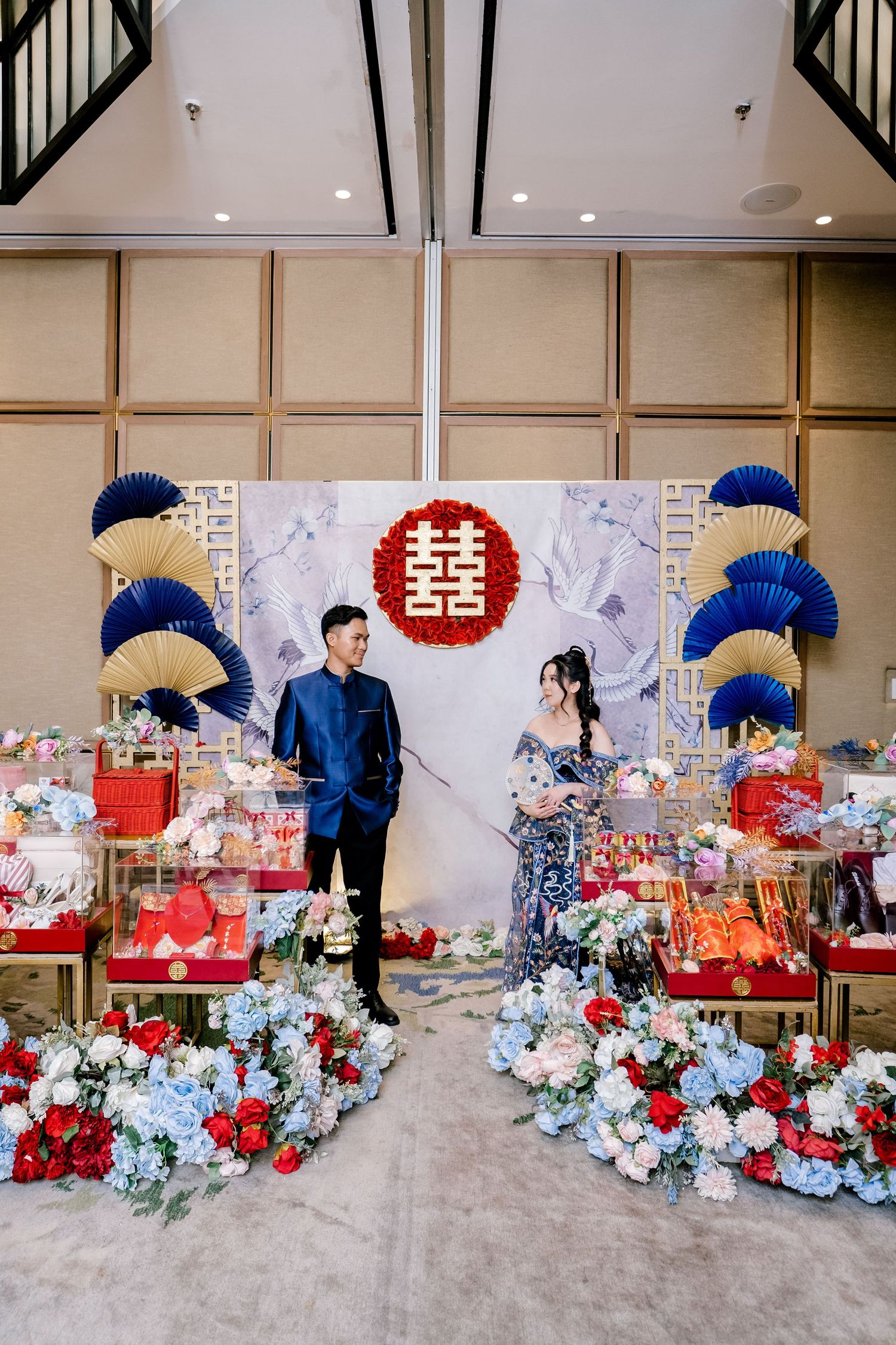 Couple poses at Chinese wedding ceremony with double happiness symbol, blue fans, red roses, and traditional gift displays.