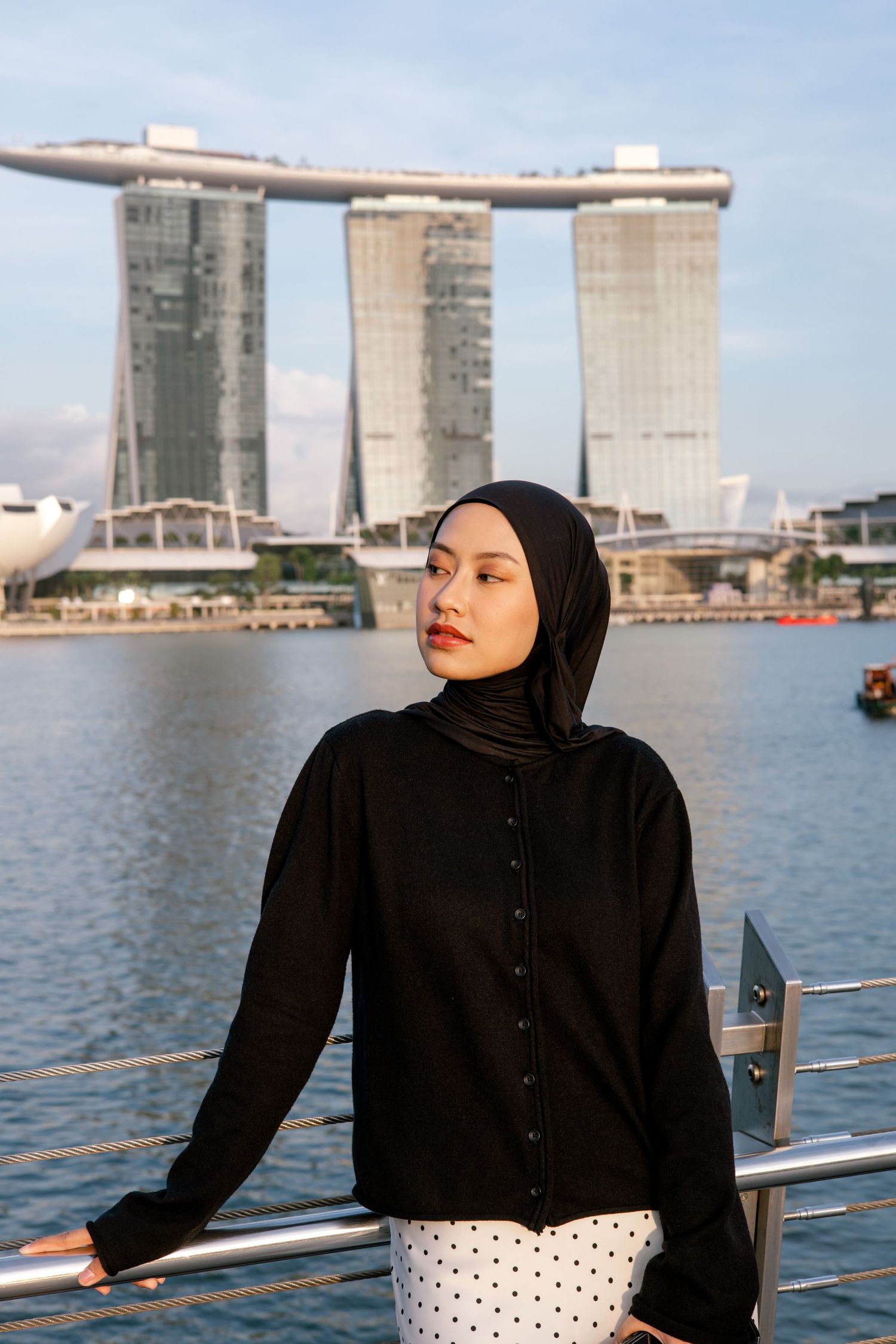 Woman in black hijab and outfit poses by waterfront railing with Marina Bay Sands hotel in background, Singapore.