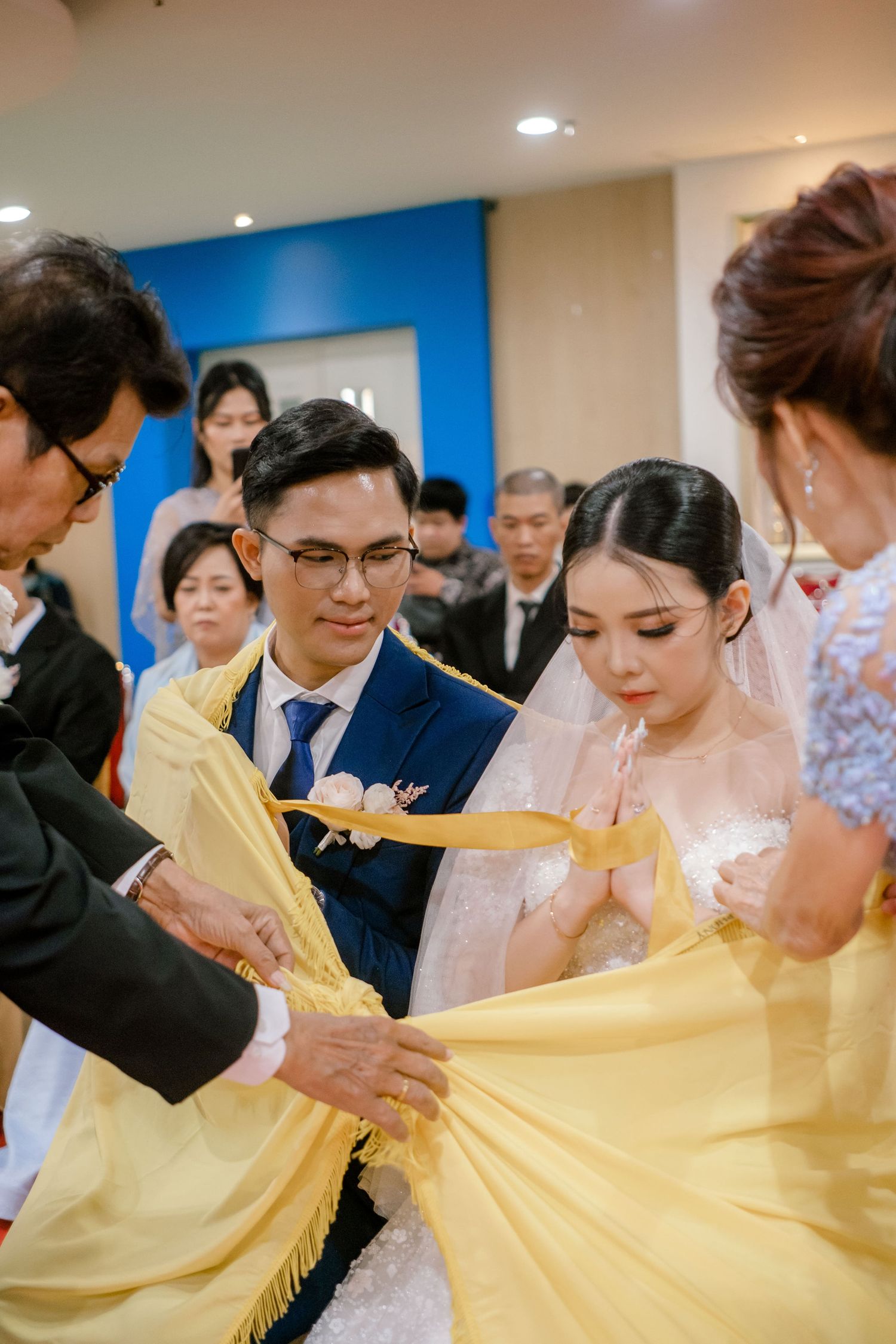 Bride and groom participate in traditional Asian wedding ceremony with yellow fabric blessing ritual, surrounded by family members.
