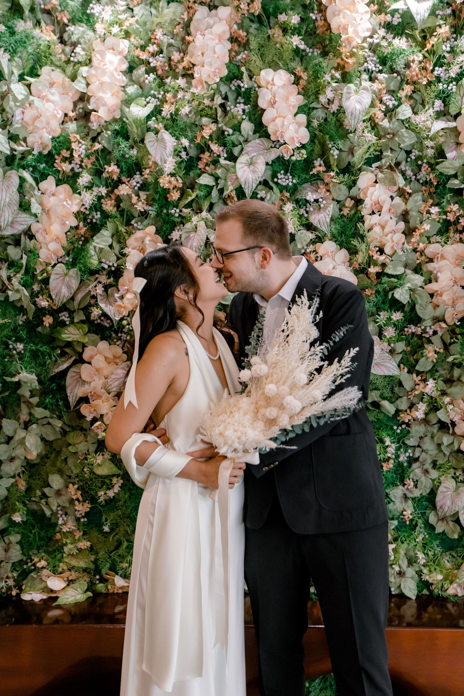 Bride and groom share a romantic moment in front of a lush floral wall backdrop at their wedding ceremony.