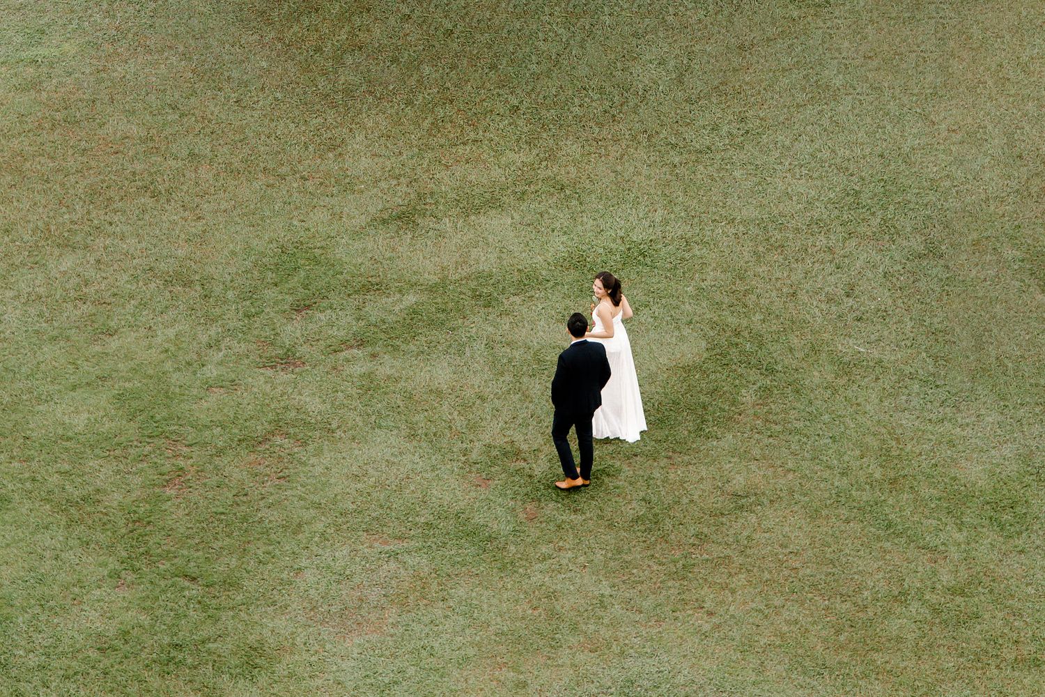 Aerial view of a bride in white gown and groom in black suit walking across a lush green grass field.