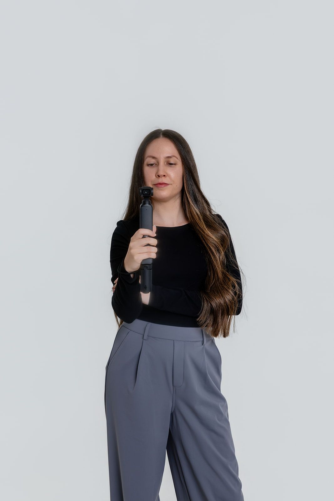 Young woman in black top and gray trousers holding a curling iron against a white background.