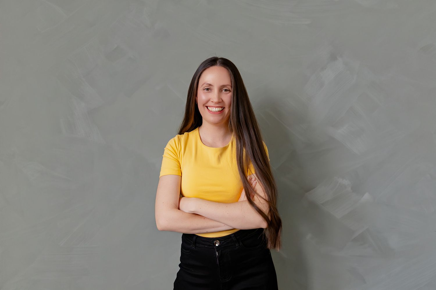 Smiling young woman with long dark hair wearing a yellow t-shirt and black skirt, arms crossed, against gray wall.