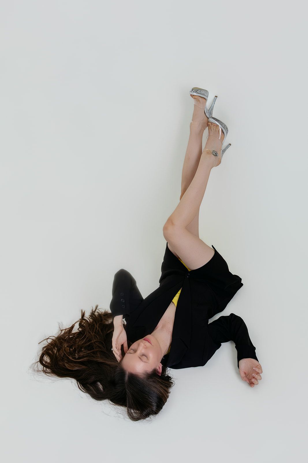 Woman in black outfit and silver heels lying on white floor with legs raised in artistic fashion pose.