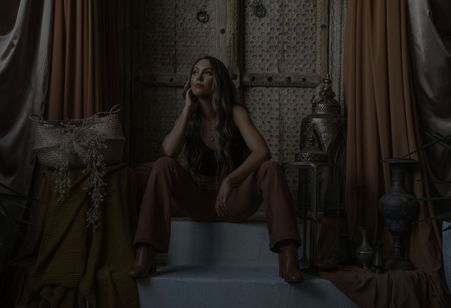 A figure sits on white steps in a dimly lit room with ornate decor and dark curtains in the background.