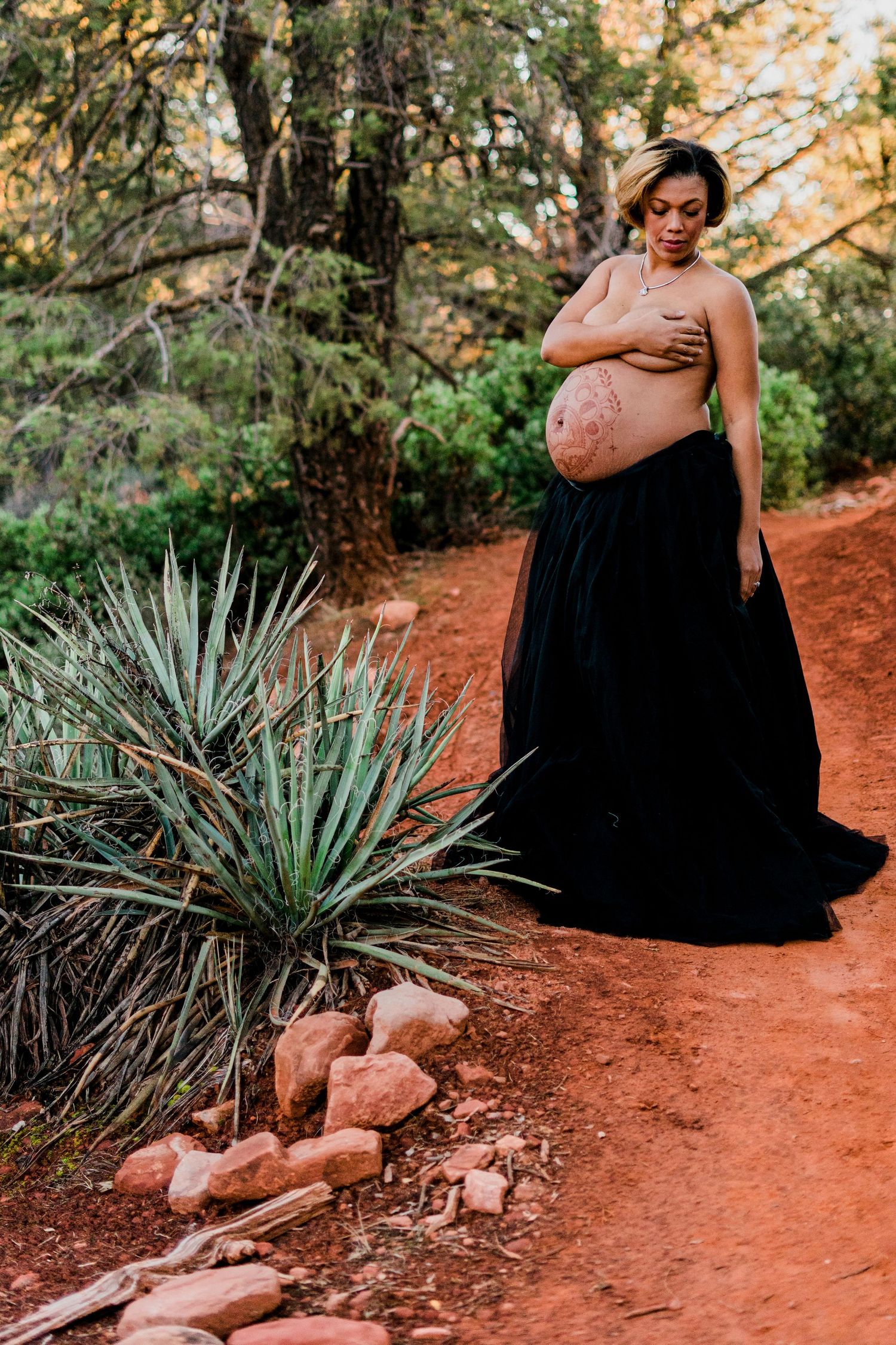 Maternity photo shoot on red dirt trail with dramatic black dress and desert plants in background.