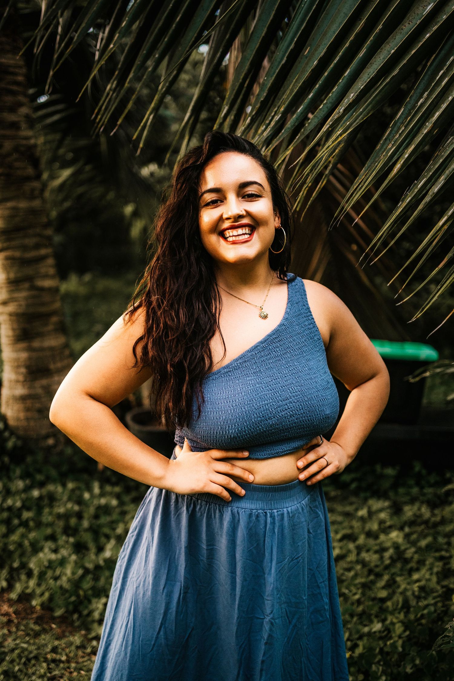 Woman in blue dress poses joyfully against tropical palm fronds background.