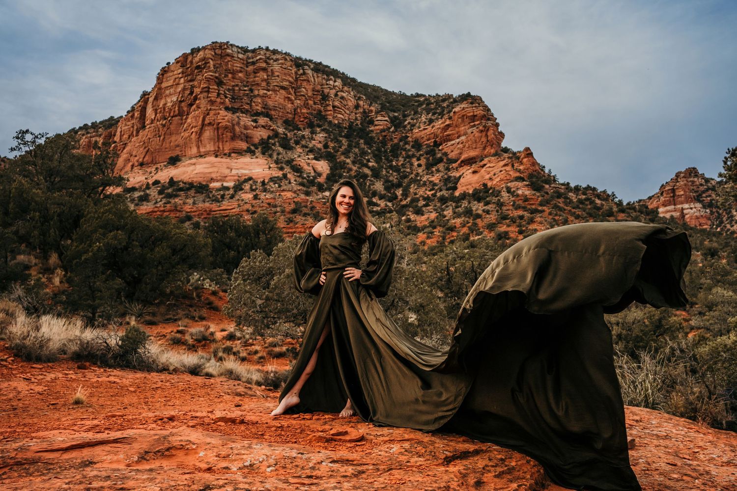 Flowing black dress billows dramatically against red rock formation in desert landscape.