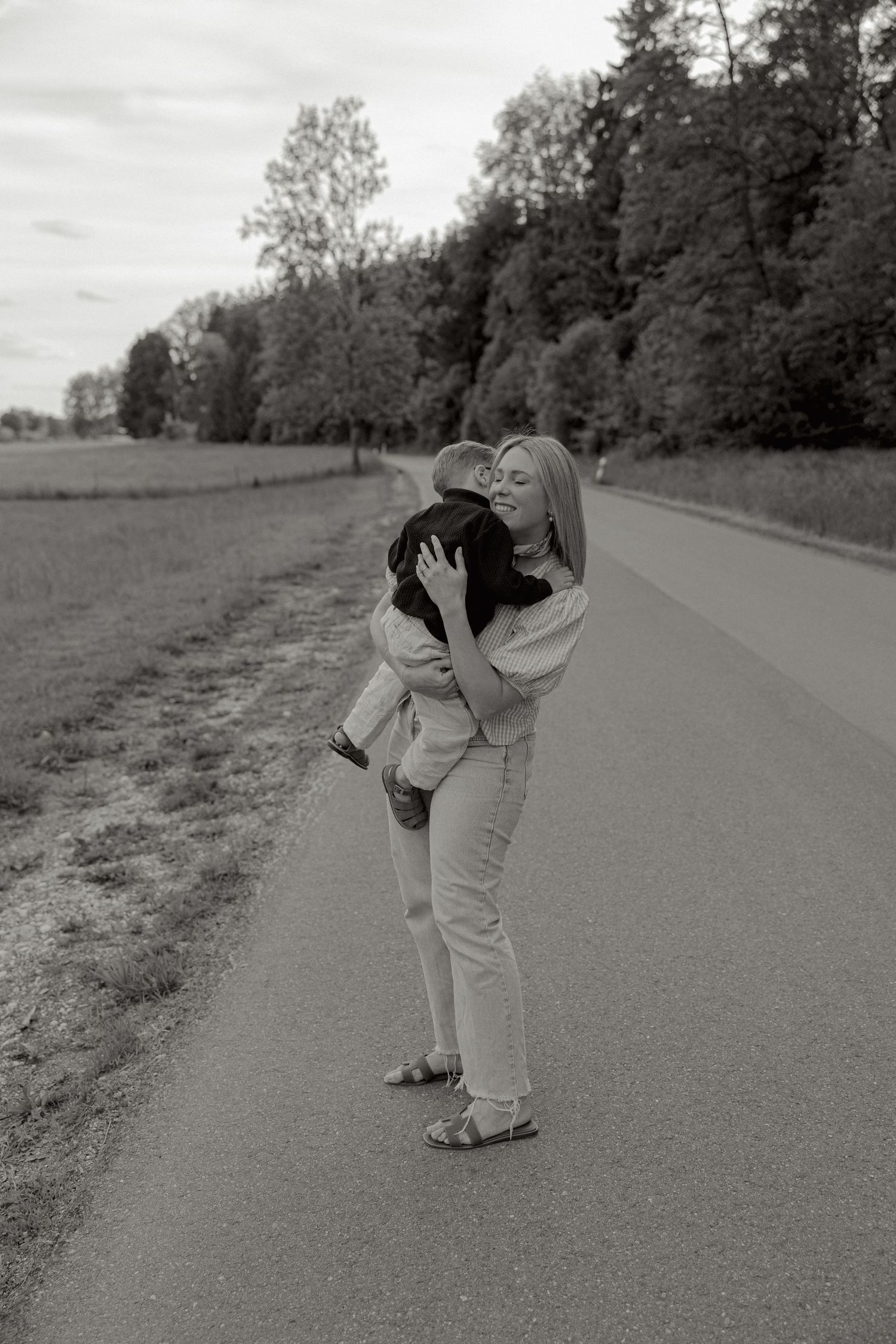 Mother holding and kissing her child on a rural road surrounded by trees, black and white.