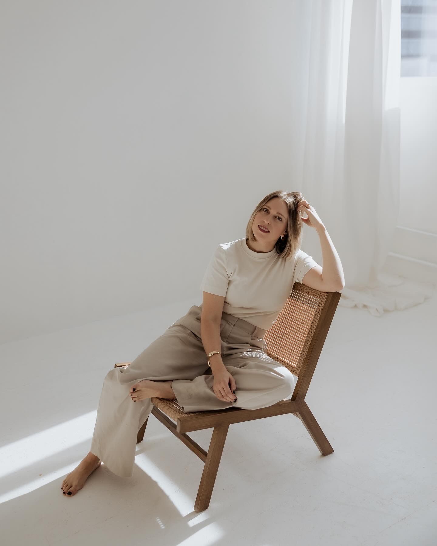 Woman sitting in a wooden cane chair in a bright white studio, wearing neutral tones.