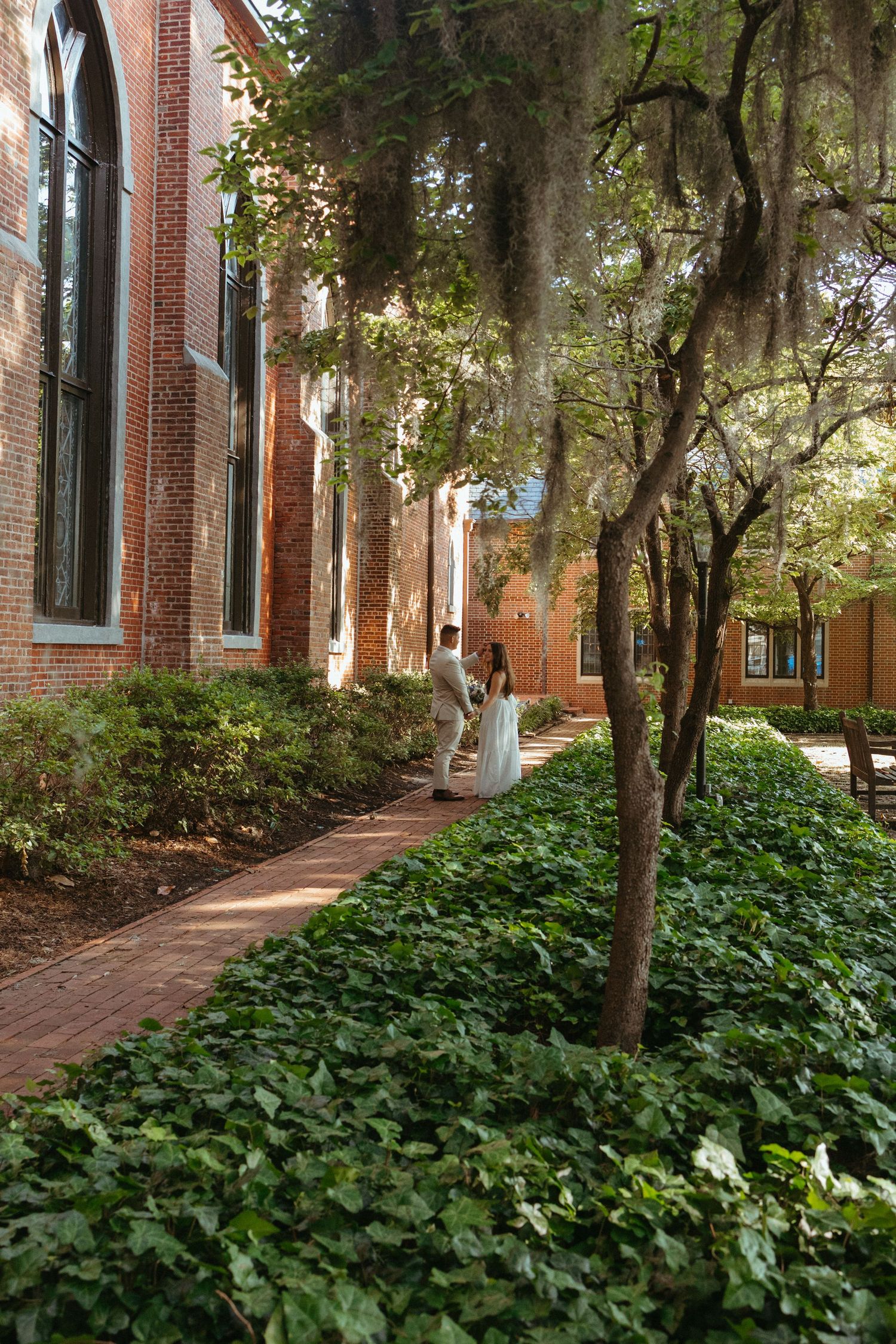 Couple in wedding attire walking along ivy-lined brick path beside historic church with Spanish moss-draped trees.