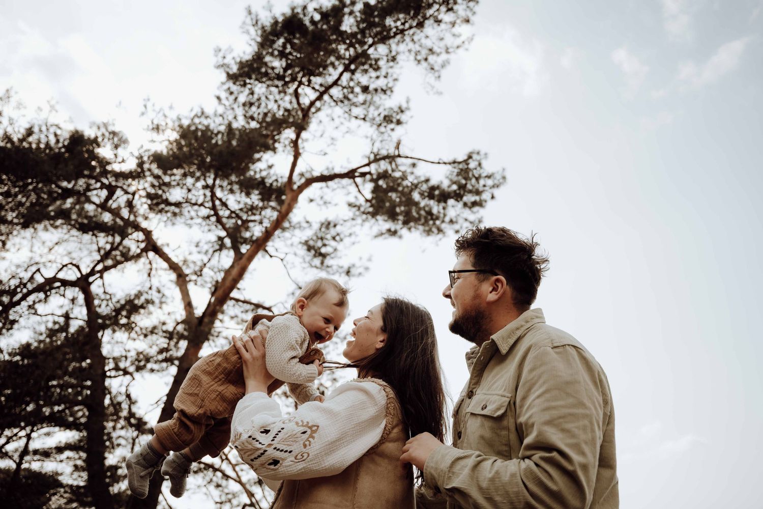 Man staat achter vrouw, vrouw houdt baby in de lucht. Op de achtergrond boomtop en lucht.
