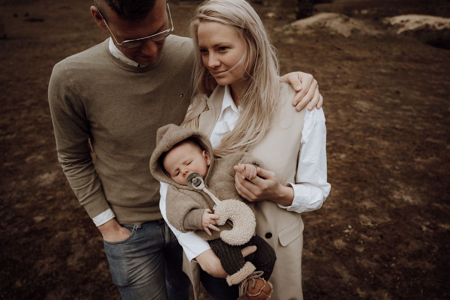 Man houdt arm om vrouw heen met hand op de schouder, vrouw heeft kind aan. Beige gekleurde kleding. Op de heide.
